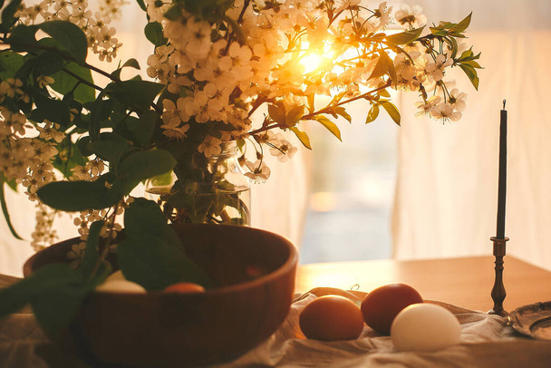 Frohe Ostern, stimmungsvoller Moment. Natürliche Ostereier und Kerze im warmen Licht des Sonnenuntergangs auf rustikalem Leinen mit Kirschblüten und grünen Blättern. Stillleben auf dem Land. Weiße und braune Eier - Foto, Bild