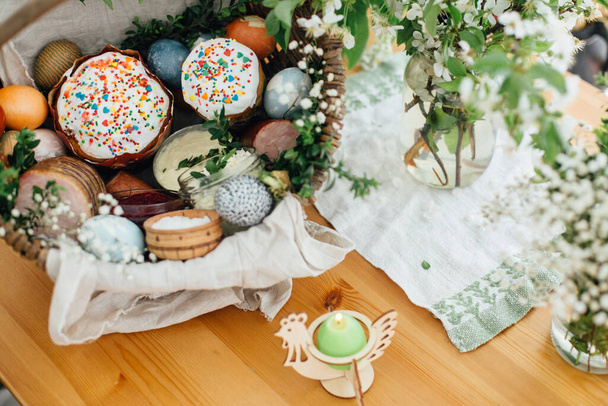 教会で祝福のための伝統的なイースターバスケット。緑のブクサスの枝やキャンドルと木製のテーブルの上の花で飾られた素朴なバスケットにイースター現代の卵、ケーキ、ハム、ビート、バター - 写真・画像