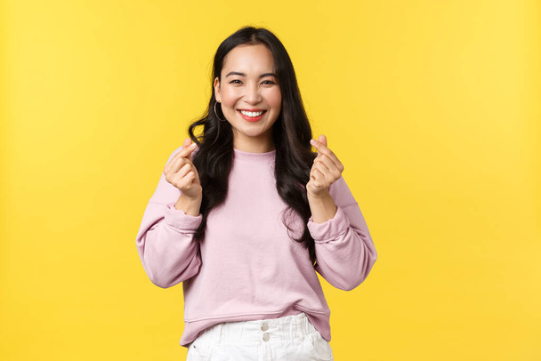 People emotions, lifestyle and fashion concept. Kawaii asian girl with beautiful smile showing korean hearts gesture sending positive and joyful vibes, standing yellow advertisement background - Zdjęcie, obraz