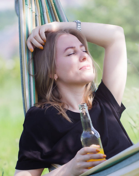 Eine glückliche Frau mit blonden Haaren entspannt und lächelt in einer Hängematte im Freien mit einer Flasche erfrischenden gelben Getränks an einem sonnigen Sommertag. - Foto, Bild