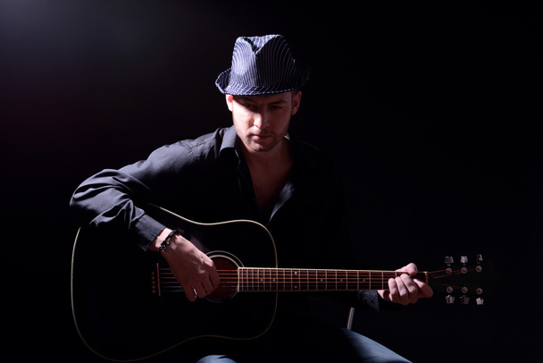 Young musician playing acoustic guitar and singing, on dark background - 写真・画像