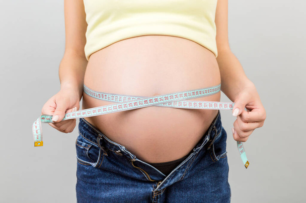 Imagen recortada de la madre embarazada en jeans desabrochados midiendo su vientre embarazada con una cinta métrica en un fondo colorido. medición de centímetros. - Foto, imagen