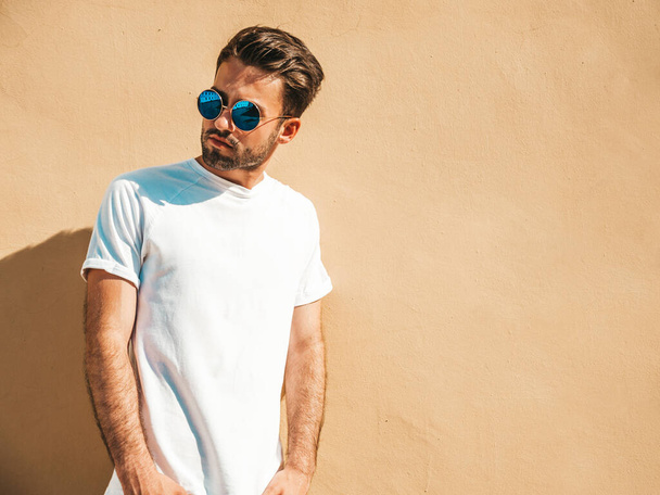 Portrait of handsome confident stylish hipster lambersexual model.Man dressed in white T-shirt. Fashion male posing in the street near wall in round sunglasses - Fotografie, Obrázek
