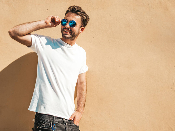 Portrait of handsome smiling stylish hipster lambersexual model.Man dressed in white T-shirt. Fashion male posing in the street near wall in round sunglasses - Foto, afbeelding