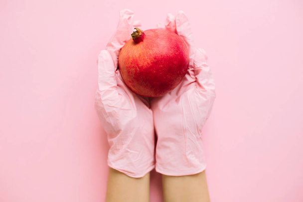 Hands in pink glove holding pomegranate on pink background flat lay. Safe shopping in quarantine. Order food online with delivery and stay home. Prevention of virus epidemic. Stay safe. - Fotó, kép