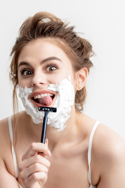 Όμορφη νεαρή λευκή γυναίκα ξυρίζει το πρόσωπό της με ξυράφι σε λευκό φόντο. Όμορφη γυναίκα με αφρό ξυρίσματος στο πρόσωπό της - Φωτογραφία, εικόνα