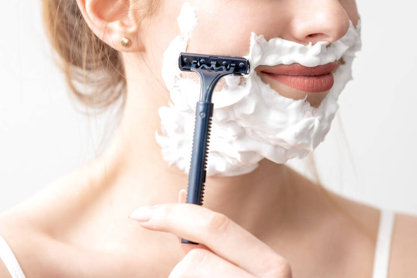 Beautiful young caucasian woman shaving her face by razor on white background. Pretty woman with shaving foam on her face - Φωτογραφία, εικόνα