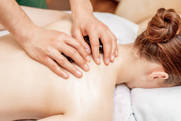 Junge Frau erhält eine Steinmassage auf dem Rücken, während die Hände des Massagetherapeuten ihr Steine auf den Rücken legen. - Foto, Bild