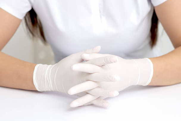 Руки в защитных белых медицинских перчатках женщины, сидящей за белым столом. Скрещенные пальцы в перчатках - Фото, изображение