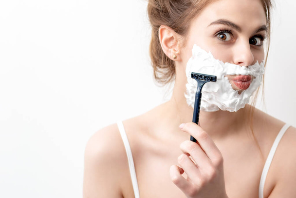 Bella giovane donna caucasica radersi il viso da rasoio su sfondo bianco. Bella donna con schiuma da barba sul viso - Foto, immagini