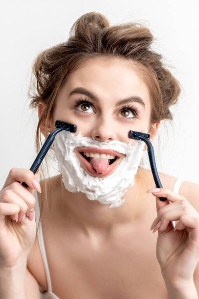 Όμορφη νεαρή καυκάσια γυναίκα ξυρίζει το πρόσωπό της από δύο ξυράφια με γλώσσα έξω σε λευκό φόντο. - Φωτογραφία, εικόνα