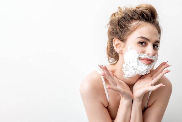 Hermosa joven mujer caucásica con espuma de afeitar en su cara posando sobre fondo blanco - Foto, Imagen