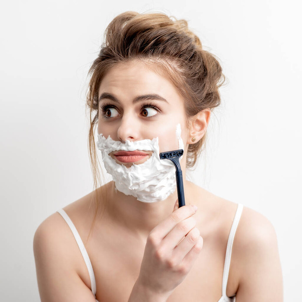 Beautiful young caucasian woman shaving her face by razor on white background. Pretty woman with shaving foam on her face - Foto, imagen