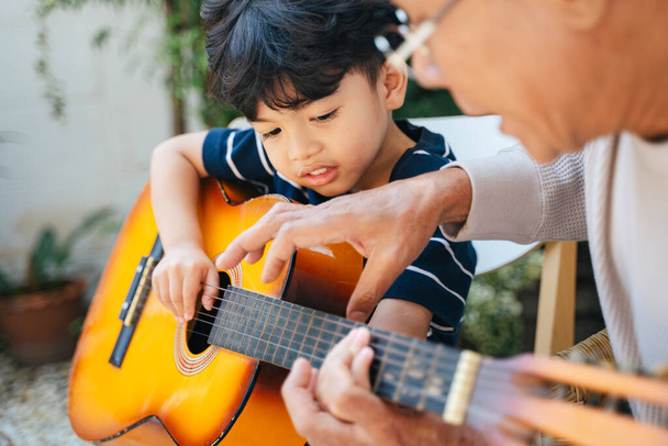 Oude man met grijs haar leert zijn kleinzoon gitaar spelen in de achtertuin van zijn huis. - Foto, afbeelding