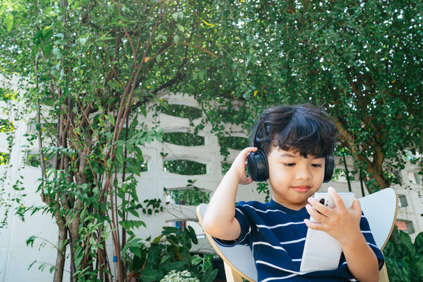 Kablosuz kulaklık takan küçük çocuk hafta sonu arka bahçede oturup akıllı telefondan çizgi filmi izliyor.. - Fotoğraf, Görsel