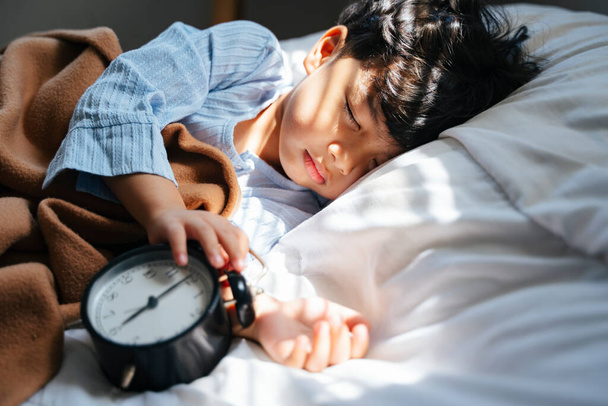 Little boy sleep at the noon on the cozy bed with sunlight from the window holding alarm clock. - Photo, Image