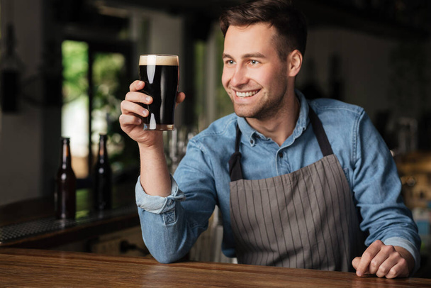 Great taste. Smiling handsome barman holds glass of dark beer in hand - Fotoğraf, Görsel