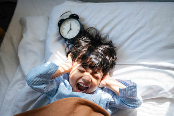 Mały chłopiec zamyka uszy, gdy dzwoni budzik, żeby go obudzić rano, ale chce więcej snu.. - Zdjęcie, obraz