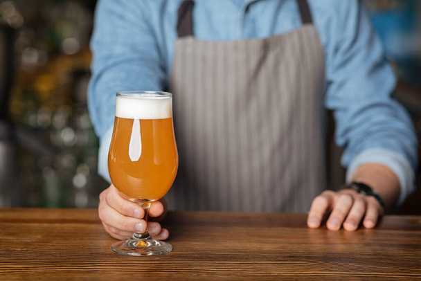 Cerveza artesanal fresca. Hombre en delantal pone en mesa de madera vidrio de cerveza ligera con espuma
 - Foto, Imagen