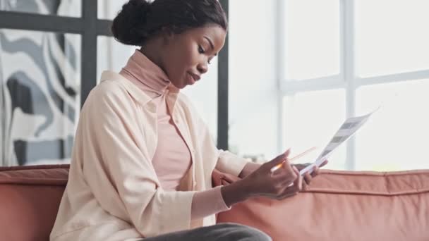 A pretty focused young african woman is choosing new colors for home renovation sitting on a couch at home - Filmmaterial, Video