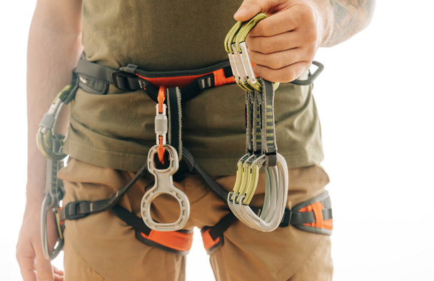 Sporty man in safety harness with climbing and mountaineering equipment holding set of quickdraws indoor on a white background. - Zdjęcie, obraz
