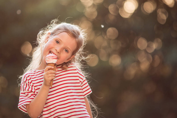 Portrait of a little girl with wavy blond hair, eating ice cream, enjoying the setting sun - Фото, изображение