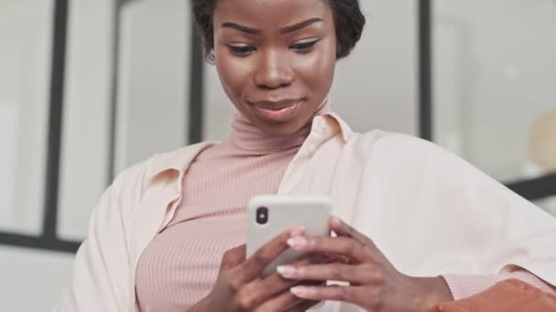 A happy pleased young african woman is using her mobile phone while sitting on a couch in the living room at home - Filmagem, Vídeo