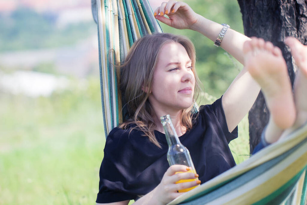 Una donna felice con i capelli biondi sta distendendosi e sorridendo in un hammock all'aperto con una bottiglia della bevanda gialla rinfrescante su un giorno d'estate soleggiato. - Foto, immagini