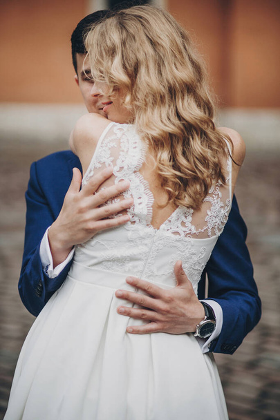 Κομψό ζευγάρι αγκαλιάζει σε ευρωπαϊκό δρόμο της πόλης. Αισθησιακή ρομαντική στιγμή. Μοντέρνα νύφη και γαμπρός ερωτευμένοι αγκαλιάζονται απαλά. Ημέρα του Αγίου Βαλεντίνου - Φωτογραφία, εικόνα