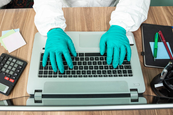 Απολύμανση άνθρωπος με ειδική λευκή σουίτα που εργάζονται σε φορητό υπολογιστή στο γραφείο top view - Φωτογραφία, εικόνα
