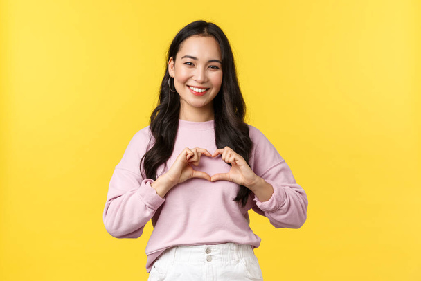 Lifestyle, Emotionen und Werbekonzept. Schöne und fürsorgliche, freundlich aussehende koreanische Mädchen breit lächelnd, mit Herzzeichen über der Brust und gelbem Hintergrund stehend - Foto, Bild