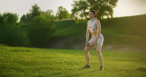 夕暮れ時、公園でダンベルを手に若い女性がしゃがみ込む。野外や草の上での訓練 - 映像、動画