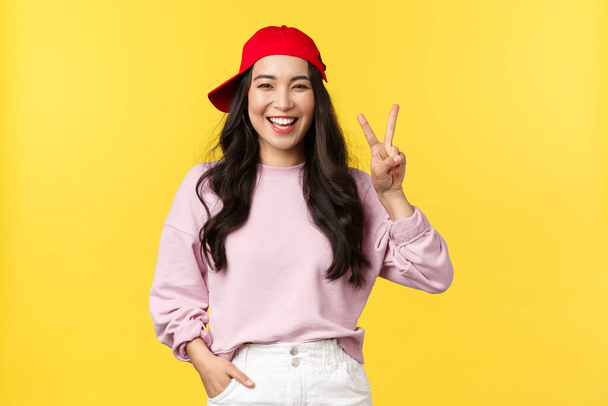 People emotions, lifestyle leisure and beauty concept. Carefree happy, asian hipster girl in summer cap, showing peace sign and smiling, enjoying sunny weekend over yellow background - Foto, afbeelding