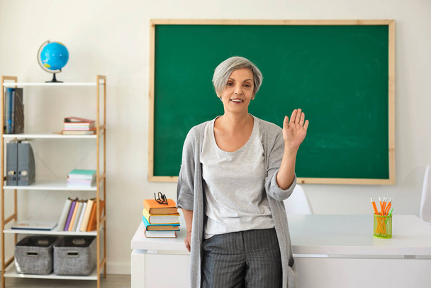 The teacher in the classroom. Woman with gray hair teacher waved her hand greeting while standing against class. - 写真・画像