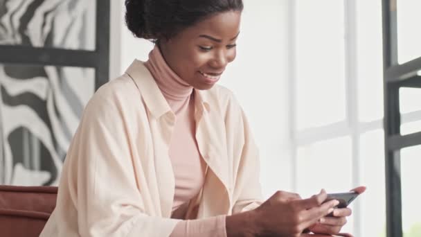Uma jovem africana sorridente linda está assistindo algo em seu telefone celular segurando-o horizontalmente enquanto trabalhava na sala de estar em casa
 - Filmagem, Vídeo