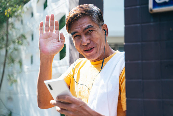 Sarı tişörtlü yaşlı adam evinin önünde duruyor ve komşusunu selamlamak için elini sallıyor.. - Fotoğraf, Görsel