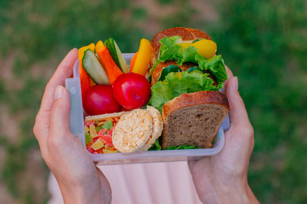Concepto de comida saludable: Joven chica hipster sosteniendo lonchera llena de sándwich, panes fritos, frutas y verduras al aire libre. Enfoque selectivo en lonchera
. - Foto, Imagen