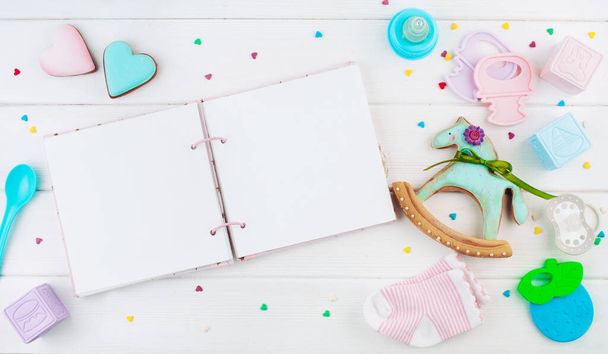 Cuaderno vacío abierto cerca de ropa de bebé y accesorios sobre fondo de madera blanca con espacio para copiar; vista superior, posición plana
 - Foto, Imagen