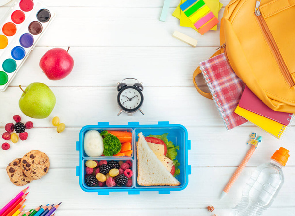 Open lunch box with sandwich, vegetables, egg, fresh berries on the white wooden background near clock and school accessories. Top view, flat lay. - Photo, Image