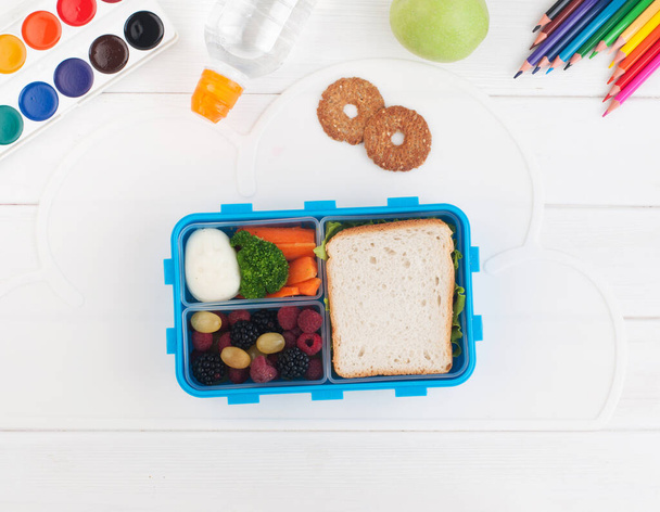 Open lunch box with sandwich, vegetables, egg, fresh berries on the white wooden background near school accessories with blank space for text. Top view, flat lay. - Photo, Image