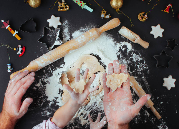 Family holiday activity. Top view of father's and kid's hands making Christmas tree cookies on black background. Flat Lay. - Photo, Image