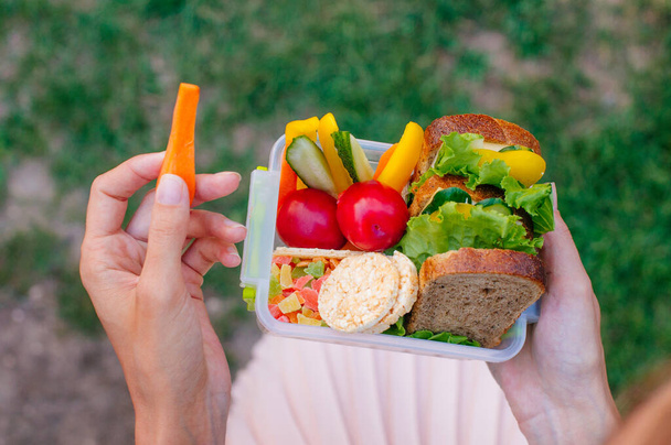 Concepto de comida saludable: Mujer joven comiendo de la lonchera llena de sándwich, panes fritos, frutas y verduras al aire libre; enfoque selectivo, vista superior
 - Foto, Imagen