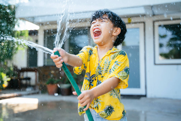 Маленький мальчик плескается водой с водяным пистолетом и резиновой трубкой у себя дома в Сонгкран день, ежегодный традиционный день Таиланда
. - Фото, изображение