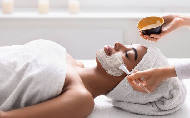 黒の女性の顔にデトックスマスクを適用する美容師 - 写真・画像