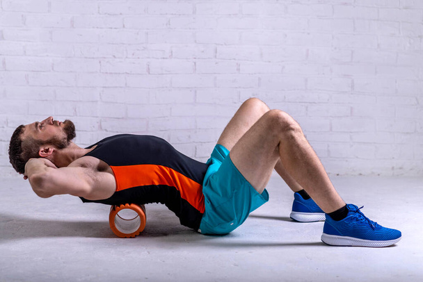 A young man in shorts is doing exercises on a foam roller on his back. Exercise on a foam roller. - 写真・画像