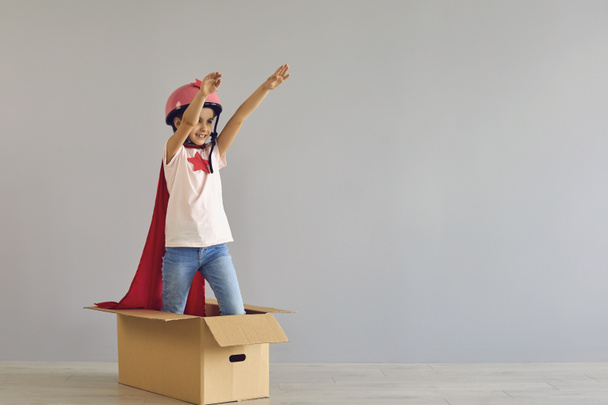 Een kind in een superheldenkostuum stak zijn hand op terwijl hij in een kartonnen doos stond met een grijze achtergrond.. - Foto, afbeelding
