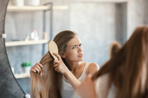 Frustrated young woman trying to brush her tangled hair in front of mirror in bathroom - 写真・画像