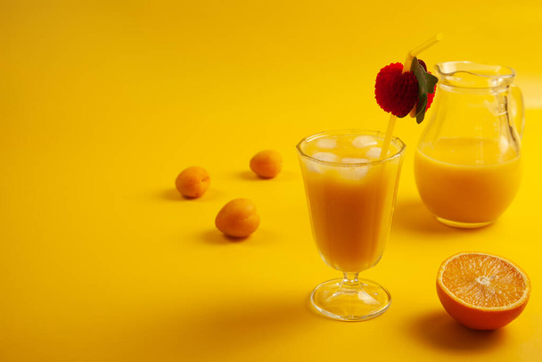 Glass with cold orange juice with ice, straw, apricots, orange half and glass jar. Yellow background. Copy space. - Foto, imagen