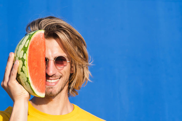 Attractive young man with long blond hair with half a watermelon covering his face on a plain blue background. Summer, sun, heat, fruit, vacation concept. - Photo, Image