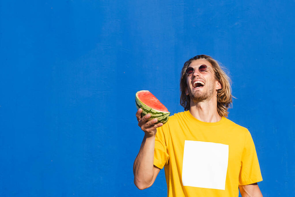 Aantrekkelijke jongeman met lang blond haar met een halve watermeloen in zijn hand glimlachend en zich vermakend op een effen blauwe achtergrond. Zomer, zon, warmte, fruit, vakantie concept. - Foto, afbeelding